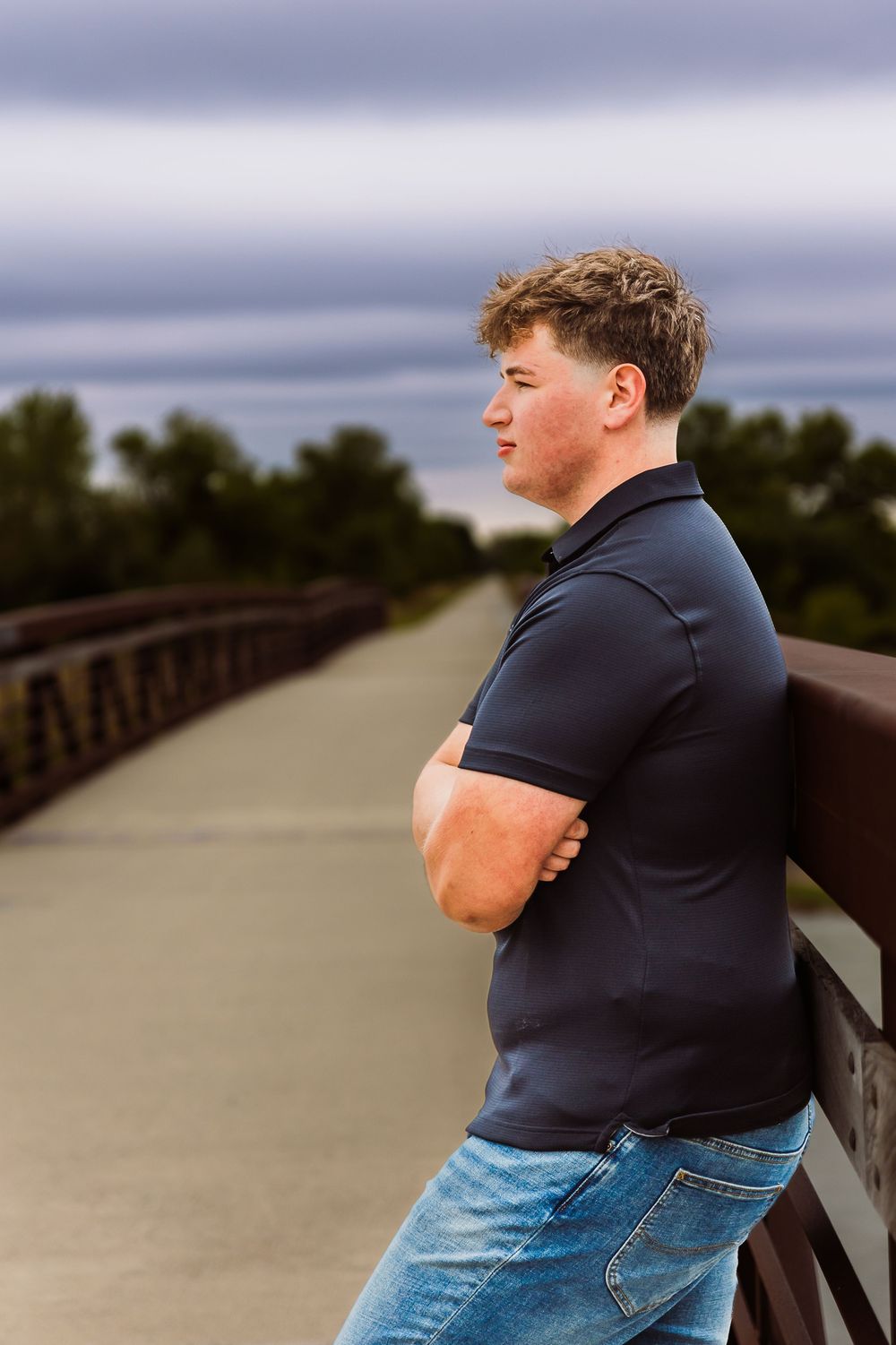 Young man in navy polo and jeans leans on bridge railing with arms crossed, gazing into distance under cloudy sky.