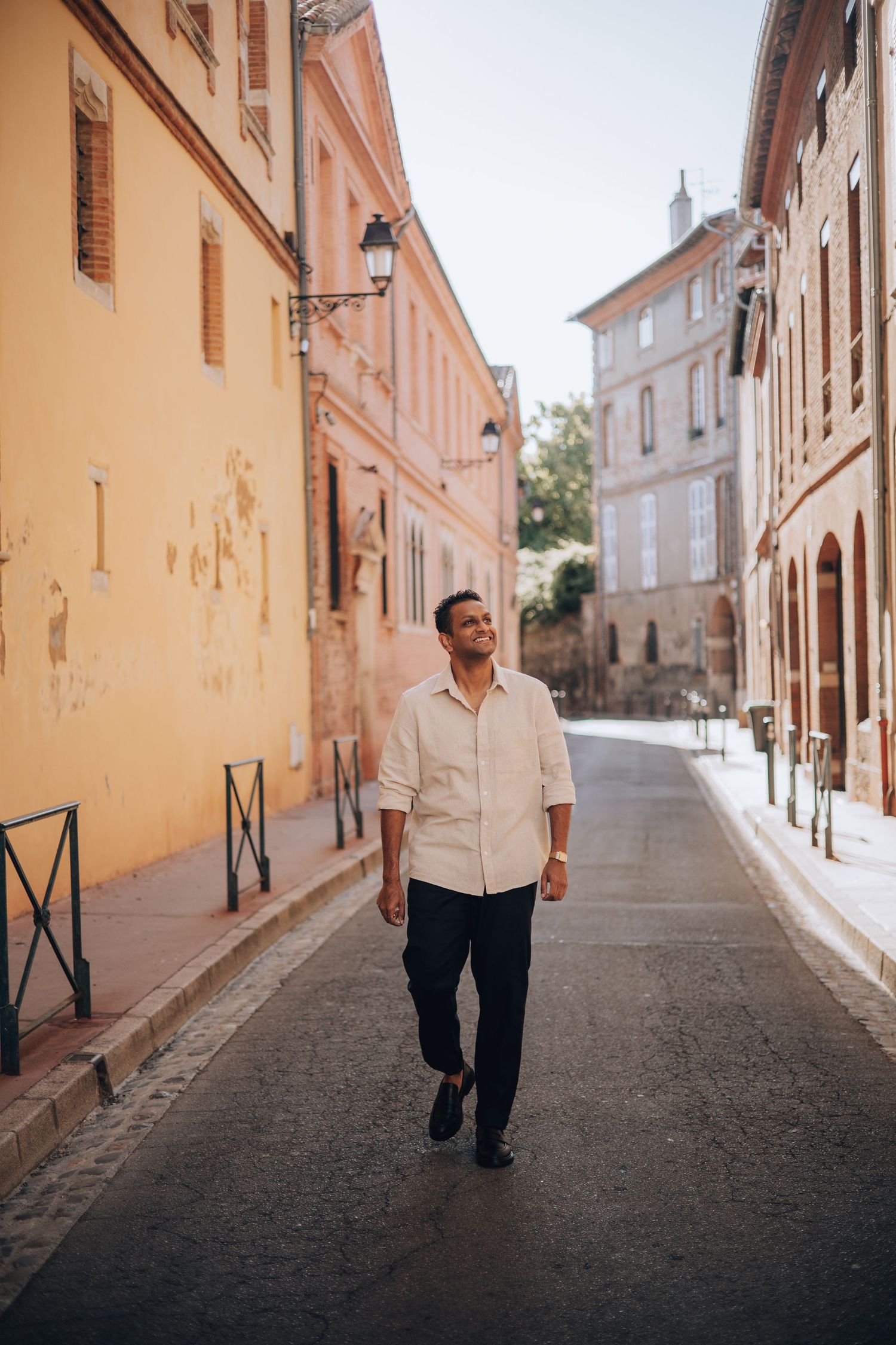 A smiling man walks down a charming European cobblestone street lined with warm-toned historic buildings on a sunny day.
