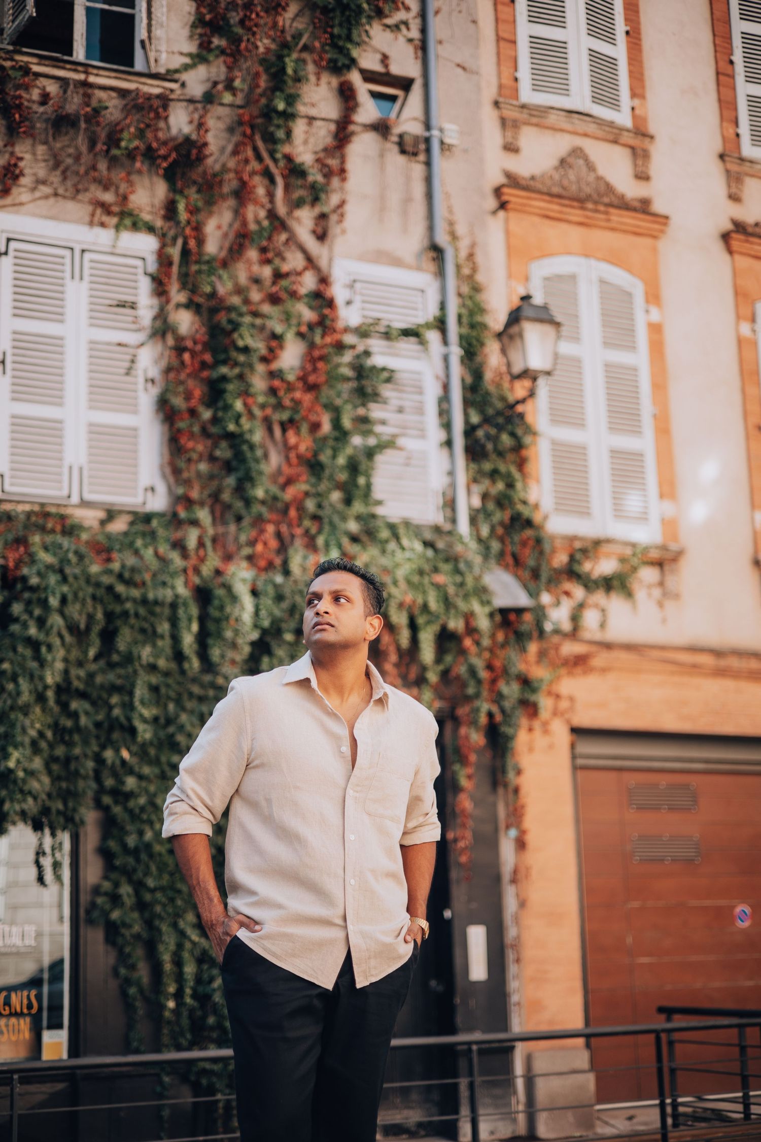 Man in white linen shirt standing outdoors near ivy-covered European building with shuttered windows.