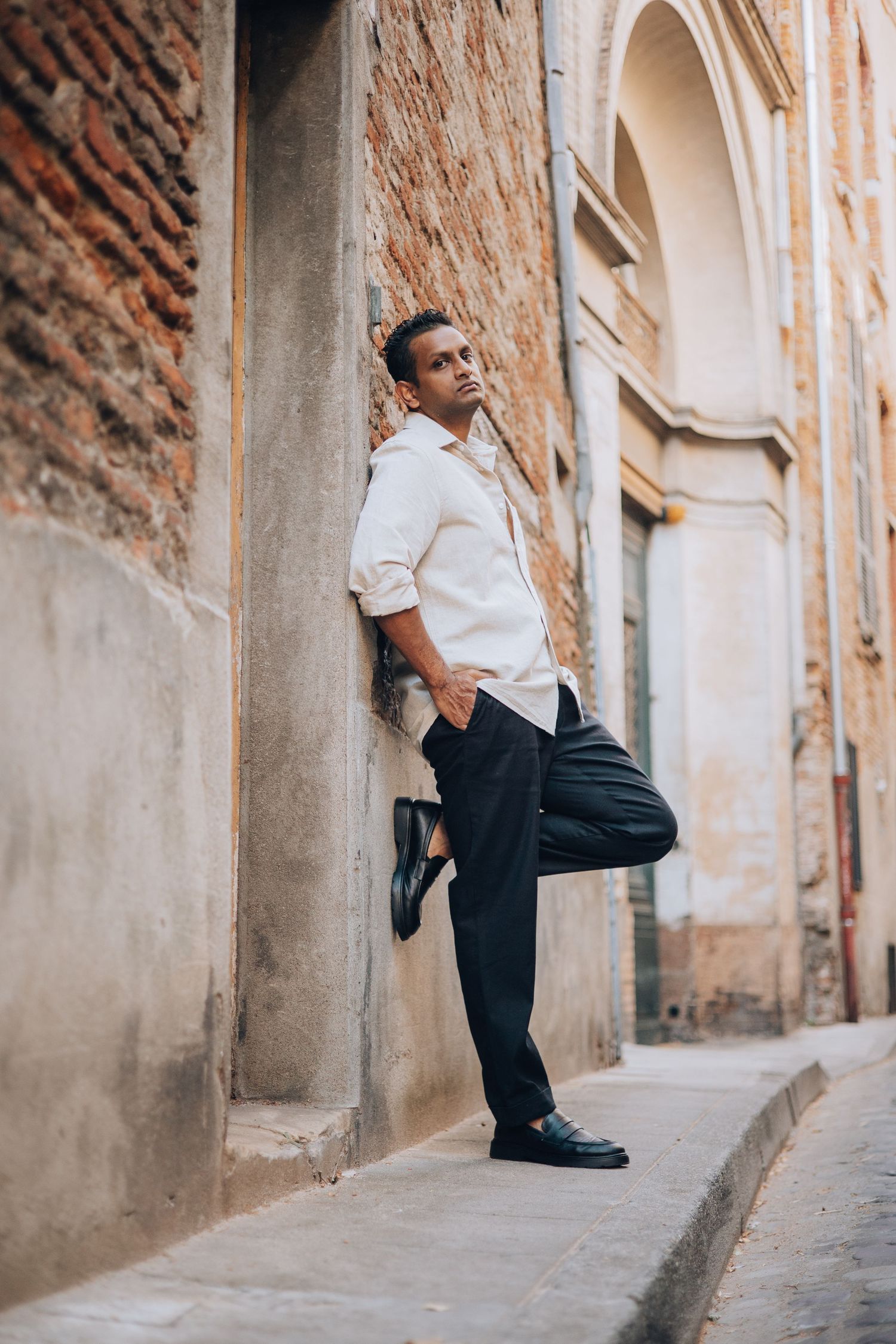 A stylish young man in white shirt and black trousers leans against an ancient brick wall in a narrow European alley.