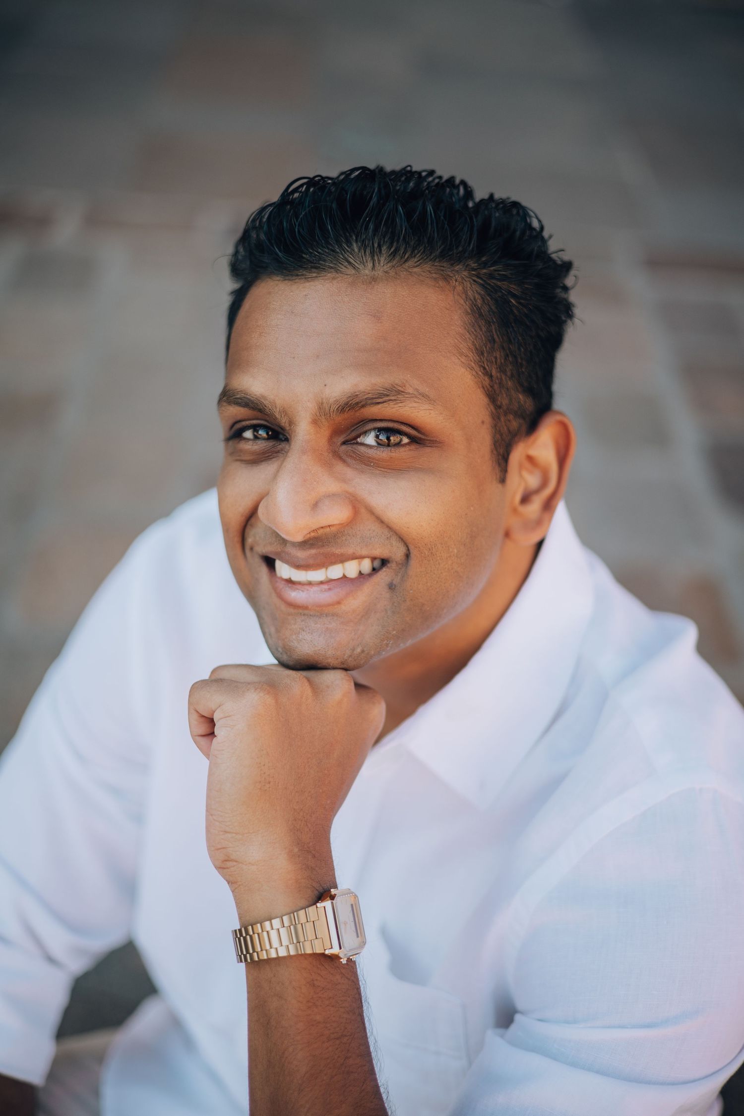 Smiling South Asian man in white shirt resting chin on hand, wearing gold watch, posed against brick wall.