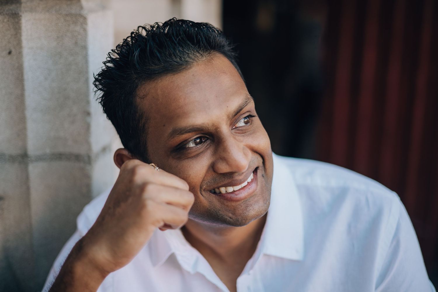Smiling South Asian man in white shirt resting chin on fist, portrait photo with soft natural lighting.