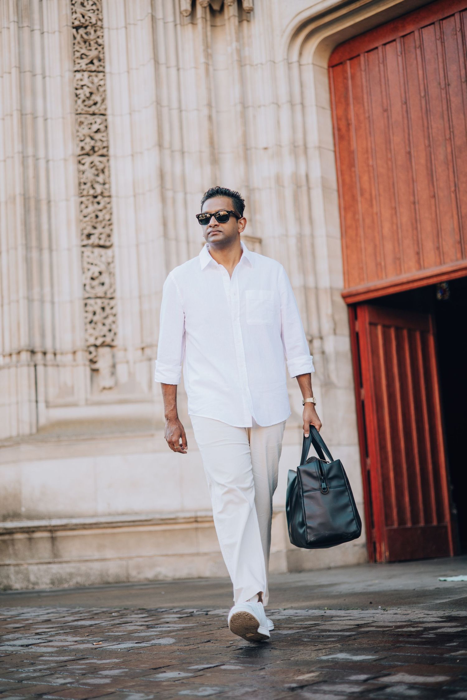 Man in white shirt and pants carrying black bag walking past ornate stone building.