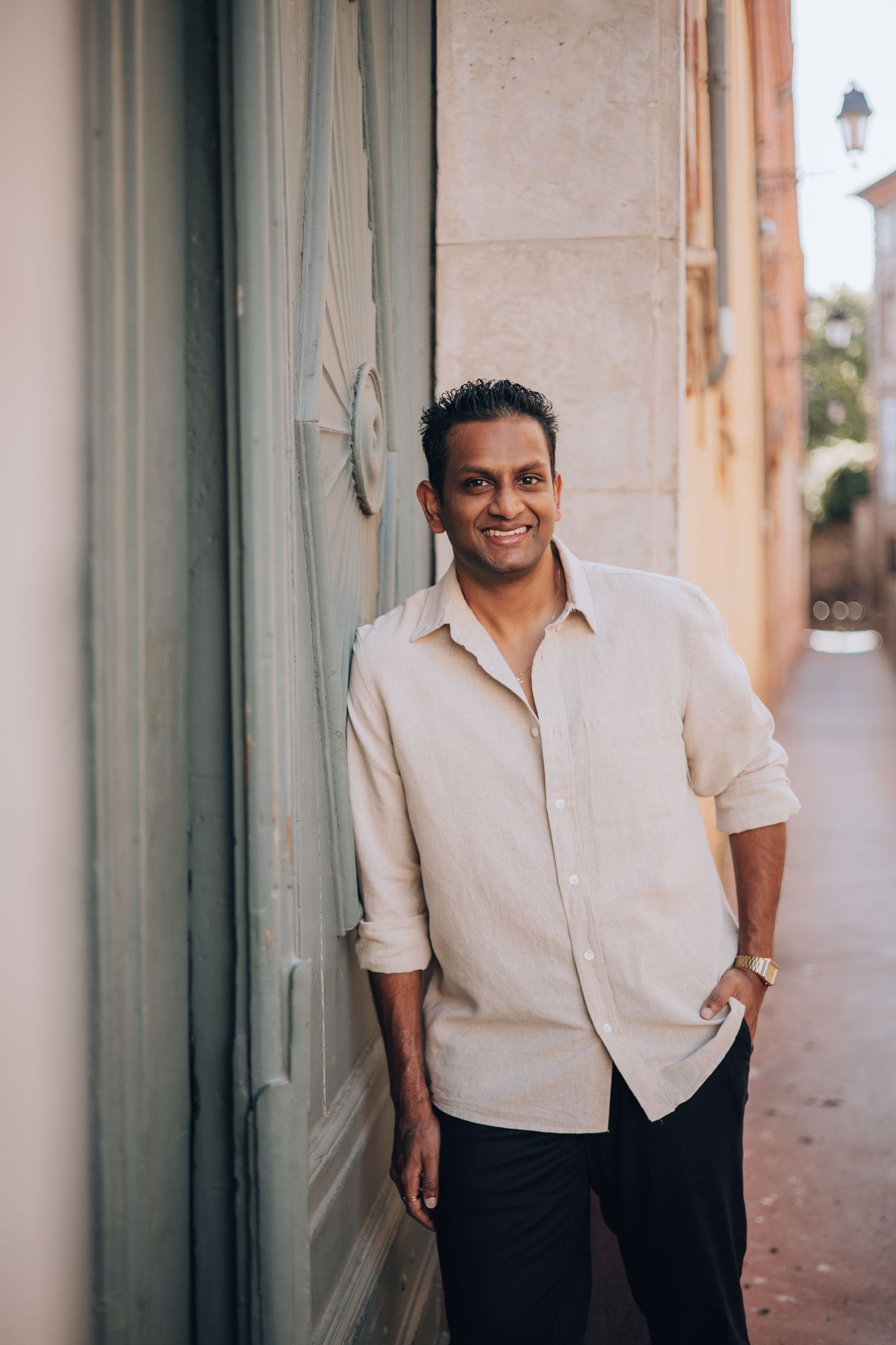Smiling man in beige linen shirt leaning against a stone wall on a sunny European street.