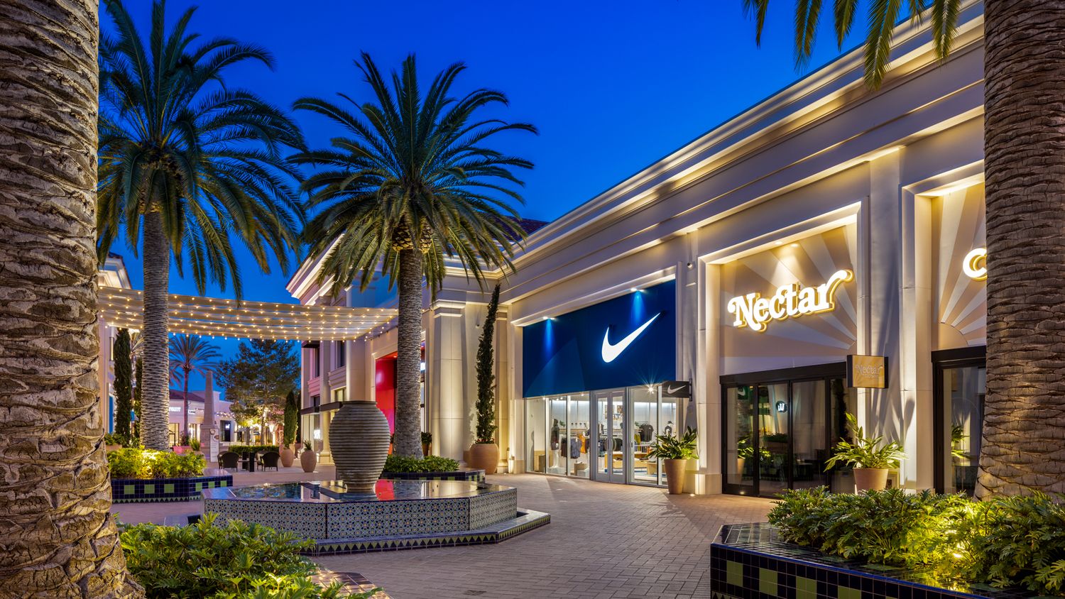 Outdoor shopping mall at dusk featuring Nike and Nectar stores, palm trees, and elegant lighting in a modern retail center.