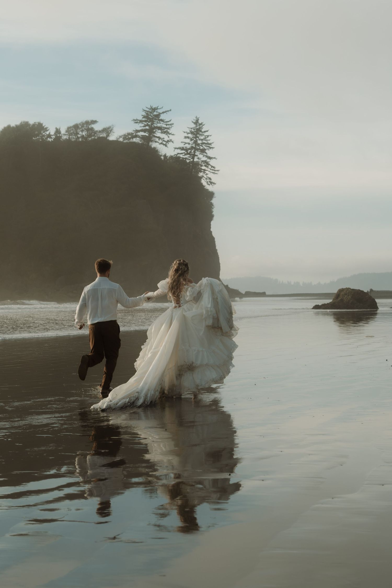 Bride and groom running on a misty beach, wedding dress flowing, with dramatic coastal cliffs and trees in the background.
