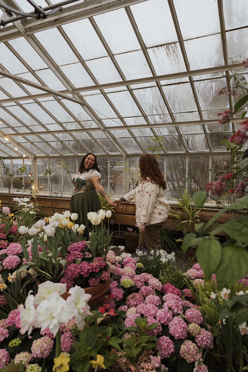 Two women smile and hold hands among pink hydrangeas and white tulips inside a glass greenhouse.