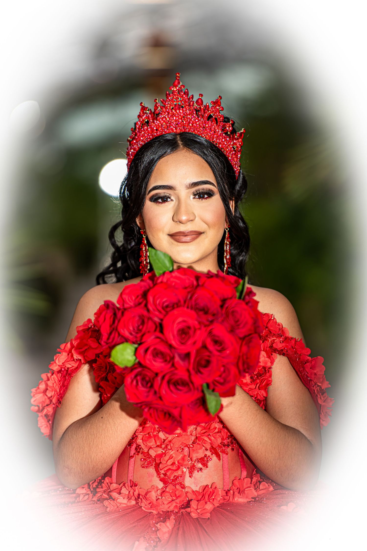Quinceañera girl wearing red crown holding bouquet of red roses in matching red floral gown.