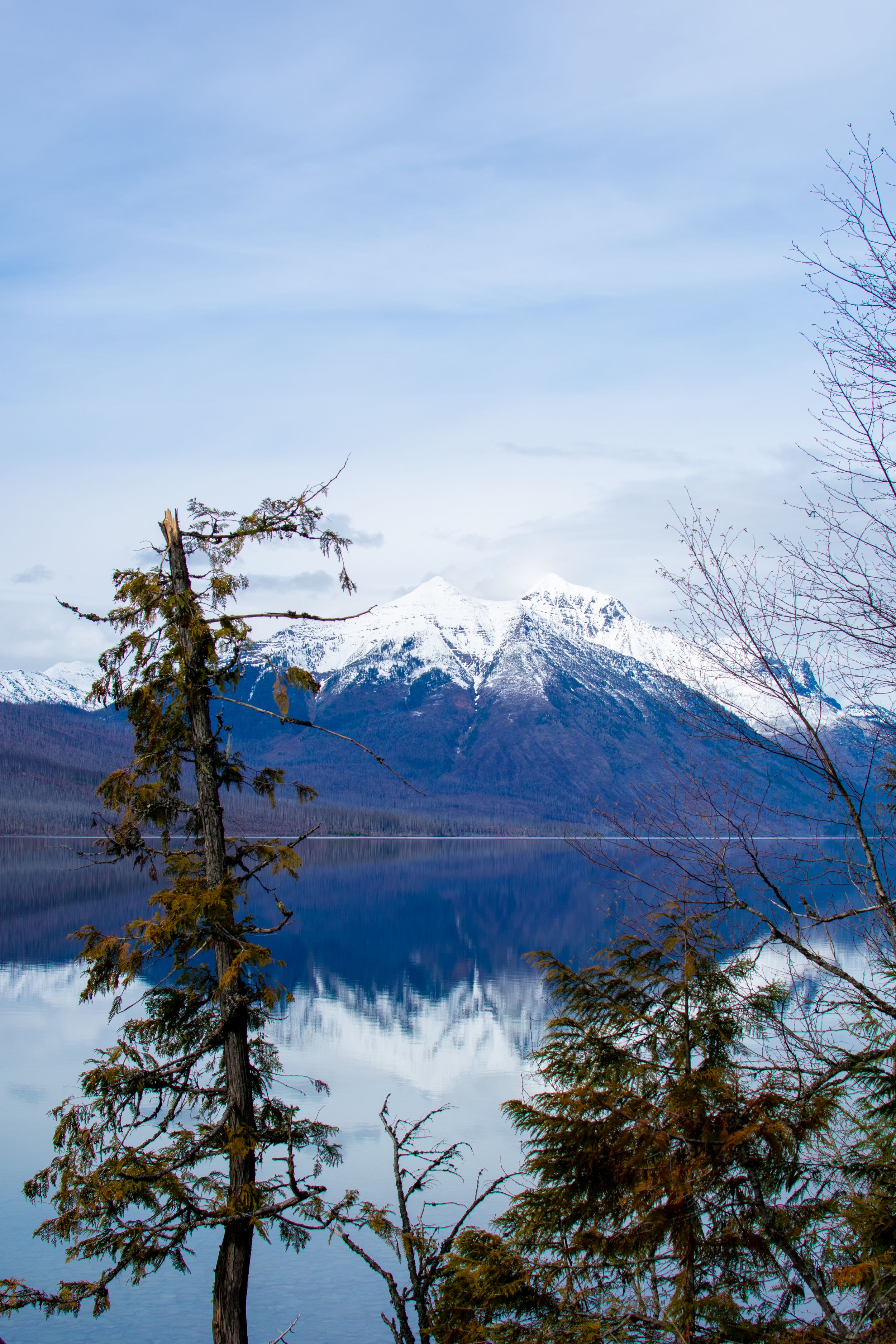 Snow-capped mountain reflects in calm blue lake framed by evergreen trees under a partly cloudy sky.