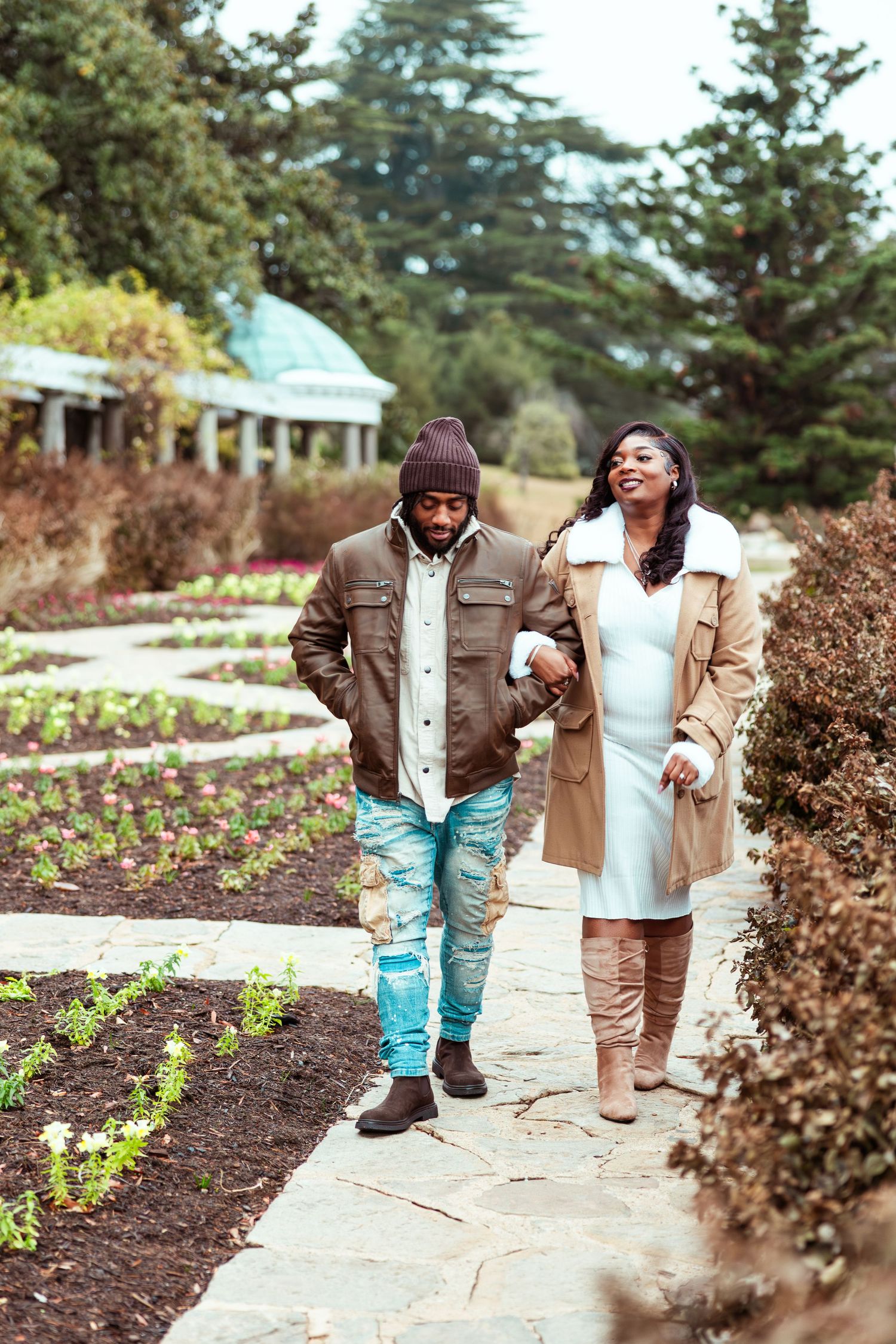 African American couple walking hand-in-hand along garden path in autumn, woman in white dress and tan coat.
