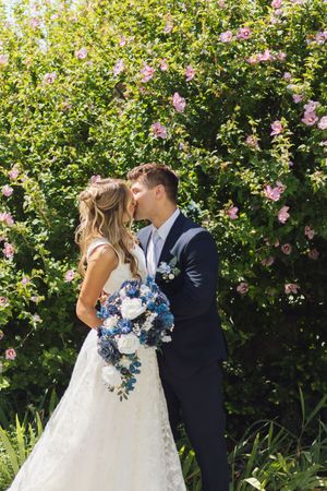 Bride and groom kissing in front of lush green bushes with pink flowers, bride holding blue and white bouquet.