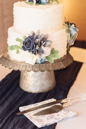Two-tier white wedding cake with navy blue roses and succulents on a rustic metal cake stand with serving utensils.