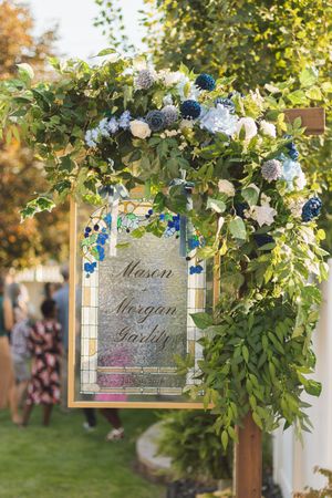 Elegant wedding sign with blue and white floral arch displaying Mason Morgan Garlity in calligraphy script outdoors.