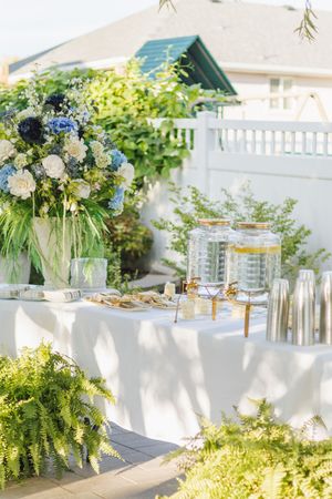Elegant outdoor wedding beverage station with blue and white floral arrangement, glass dispensers, and white tablecloth.
