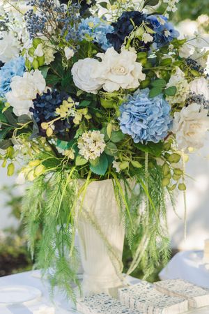 Elegant floral arrangement featuring white roses, blue hydrangeas, and lush greenery in a white vase on a decorated table.