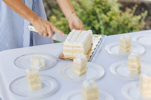 Person slicing a white layered cake onto clear plates at an outdoor event on a white table.