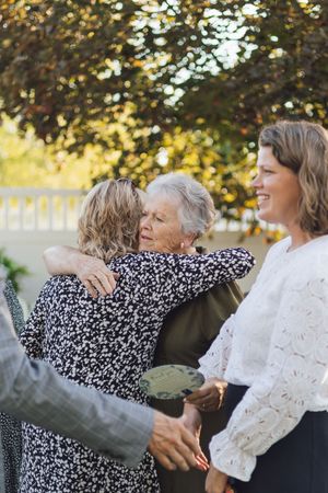Elderly woman hugging a younger woman outdoors at a family gathering, with another smiling woman standing nearby.
