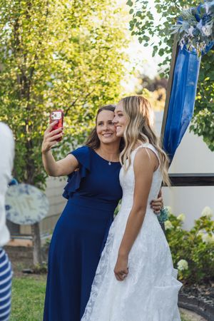 Bride in white lace gown takes selfie with bridesmaid in navy blue dress at outdoor wedding ceremony.