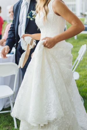 Bride in white lace wedding gown holding her dress while standing on green grass.