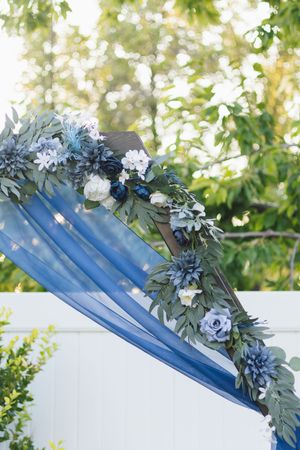 Blue floral wedding arch with roses, greenery, and draped navy chiffon fabric outdoors.