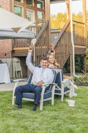Bride and groom celebrating outdoors, holding up shoes while seated in a chair on a green lawn at a backyard wedding reception.