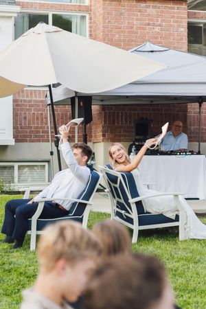 Bride and groom laughing and waving programs at outdoor wedding reception with DJ and patio umbrella.
