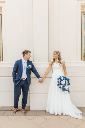 Bride and groom holding hands in blue suit and white lace wedding dress with blue bouquet.