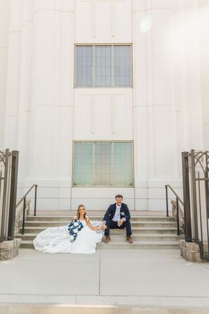Bride and groom sit on steps of white building holding blue bouquet in elegant wedding portrait.