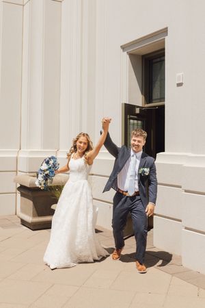 Joyful newlyweds raising hands outside white building, bride in lace gown holding blue bouquet.