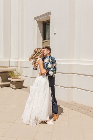 Bride and groom embracing and smiling outside a white building on their wedding day, groom holding a floral bouquet.