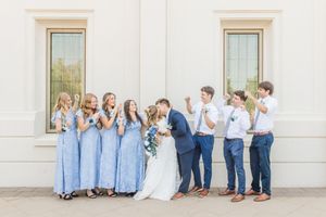 Wedding party poses outside a white building, bridesmaids in blue dresses and groomsmen in navy suits laughing as couple kisses.
