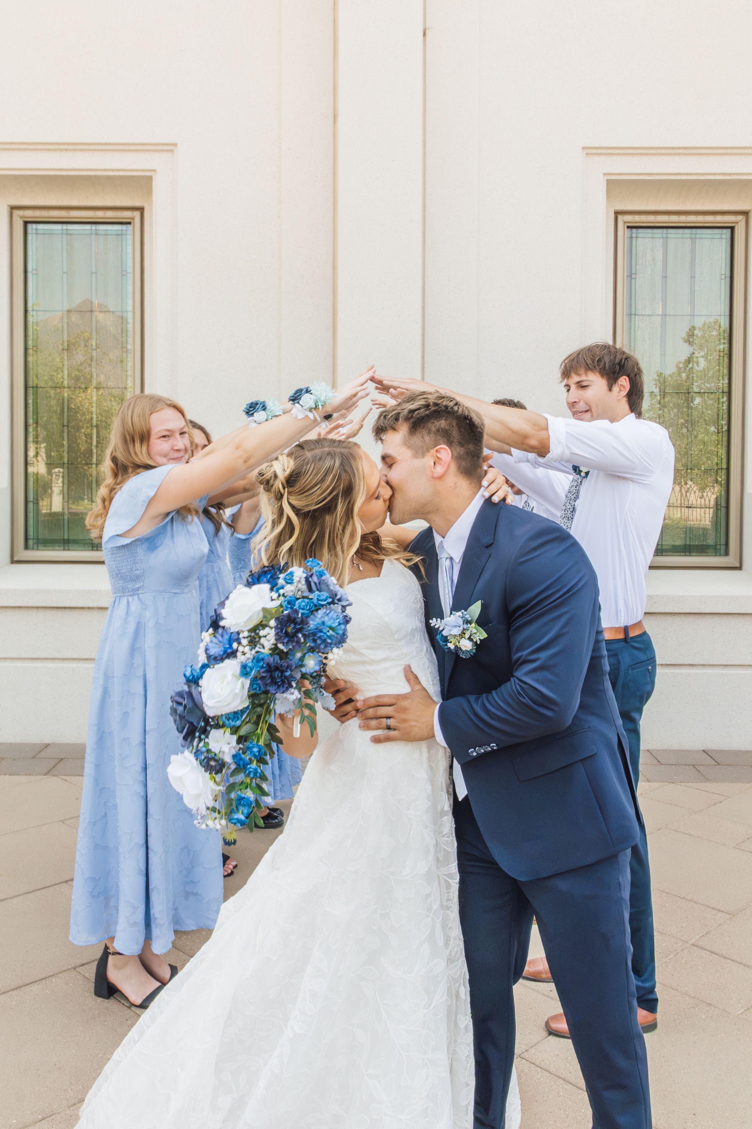 Bride and groom share a kiss while wedding party members hold flowers above them outside a white building.