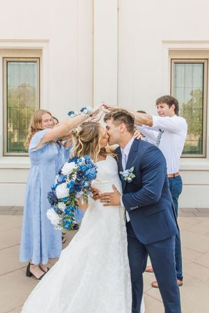Bride and groom share a kiss while wedding party members hold flowers above them outside a white building.