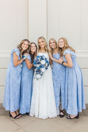 Bride in white lace gown holds blue and white bouquet surrounded by four bridesmaids in light blue dresses.