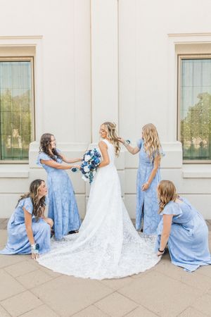 Bride in white lace gown with four bridesmaids in blue dresses arranging her train and veil outdoors.