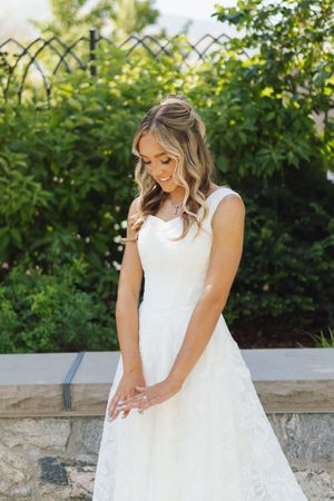 Bride in white lace A-line gown smiling downward, standing outdoors with lush green hedges in background.