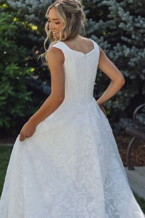 Elegant bride in a white floral lace ball gown with cap sleeves, posing outdoors among lush green trees.