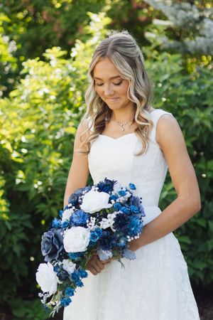 Bride in white lace dress holding blue and white floral bouquet, smiling outdoors with green foliage background.