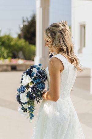 Bride in white lace dress holding cascading blue and white floral bouquet, standing outdoors near white columns.