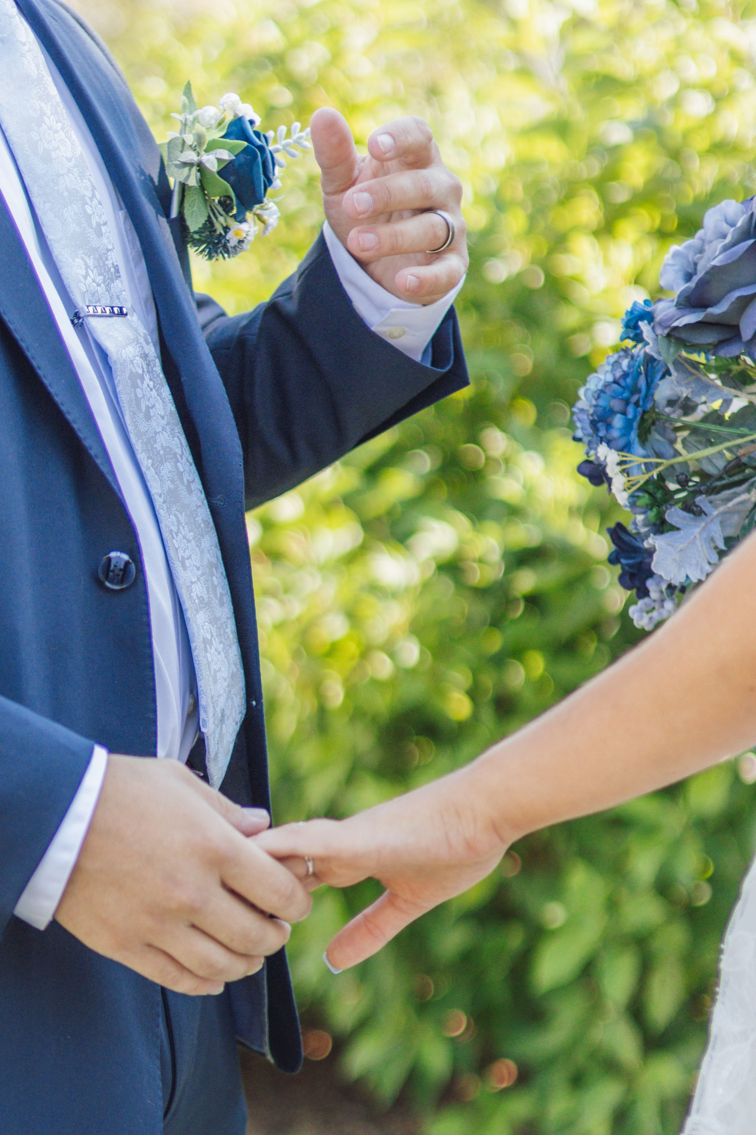 Bride and groom exchanging rings outdoors with blue boutonniere and floral bouquet in garden setting.