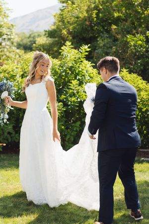 Groom helping bride with her white lace wedding dress train on a sunny green lawn with lush garden background.