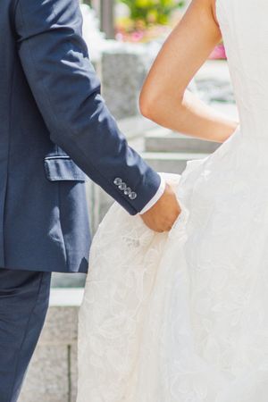 Bride and groom holding hands, wearing white lace wedding dress and navy blue suit, standing outdoors.