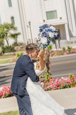 Bride and groom sharing a romantic kiss outdoors with blue and white wedding bouquet.