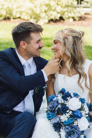 Smiling groom tenderly touches bride's chin as they sit together outdoors, bride holding blue and white bouquet.