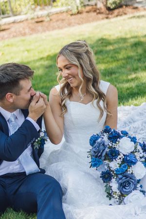 Groom in navy suit kissing bride's hand while she smiles, holding blue and white bouquet on grass.