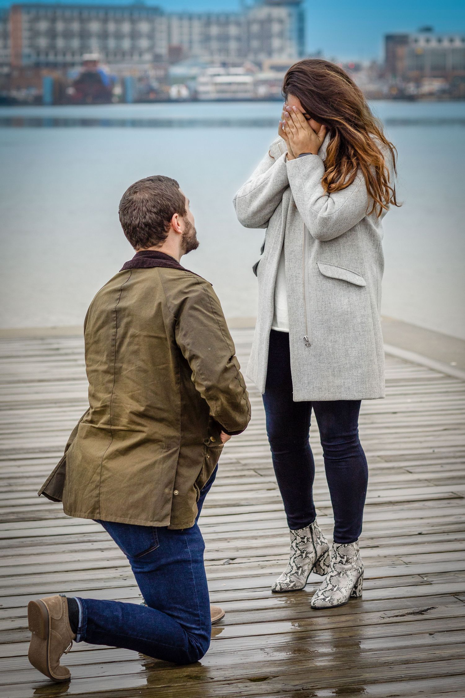Man proposes on one knee to surprised woman covering her face on a waterfront dock.