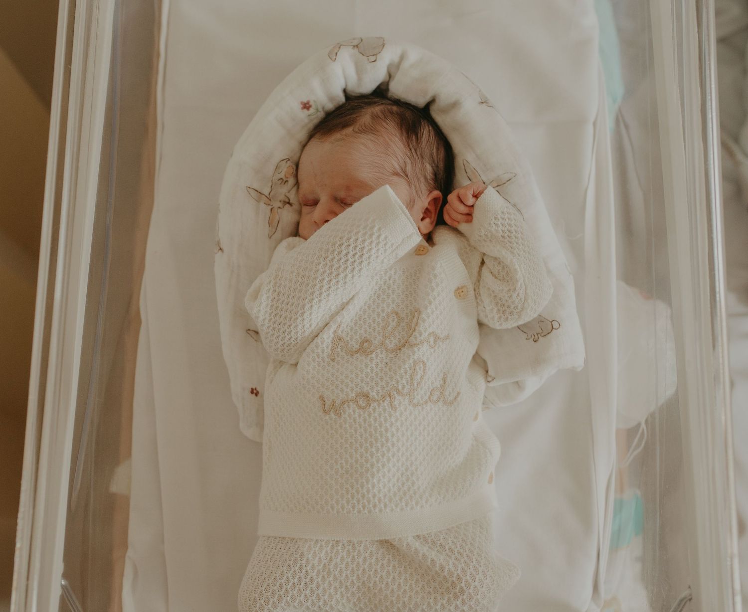 Newborn baby wrapped in a white 'hello world' knit swaddle blanket, sleeping peacefully in a hospital bassinet.