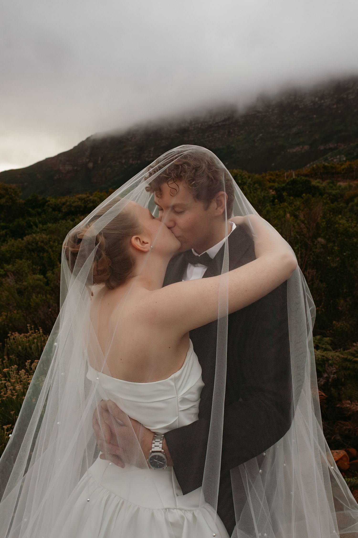 Bride and groom kissing under flowing veil with dramatic misty mountain landscape behind them.