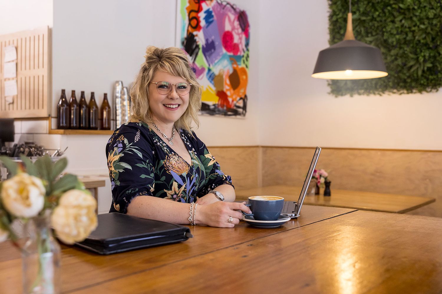 Catherine Doocey from Border Leadership smiling at her laptop with coffee in a bright modern café, colourful artwork on wall behind her.