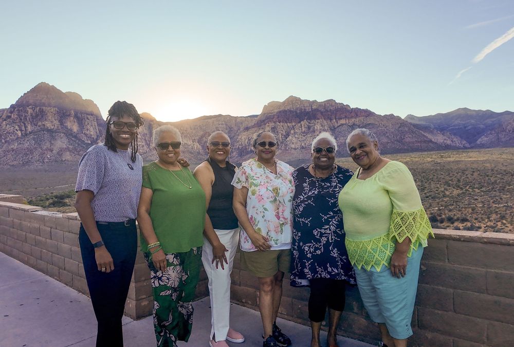 Seven women pose together at Red Rock Canyon overlook with stunning desert mountains and sunset sky behind them.
